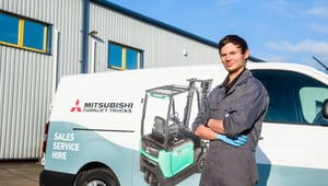 Ein Servicetechniker steht in Arbeitskleidung mit verschränkten Armen vor einem Firmenfahrzeug von Mitsubishi Forklift Trucks.