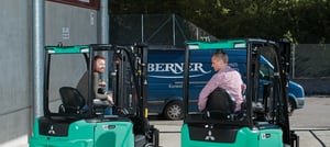 Zwei Männer sitzen einander zugewandt auf türkisfarbenen Gabelstaplern auf einem Industriegelände; im Hintergrund steht ein blauer Lieferwagen