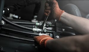 Hände einer Person mit Handschuhen, die mit einem Schraubenschlüssel an Hydraulikschläuchen arbeitet.