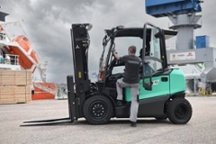 Ein Mann steigt in einen türkisfarbenen Gabelstapler ein, der auf einem Industriehof zwischen Holzkisten und Kränen steht.